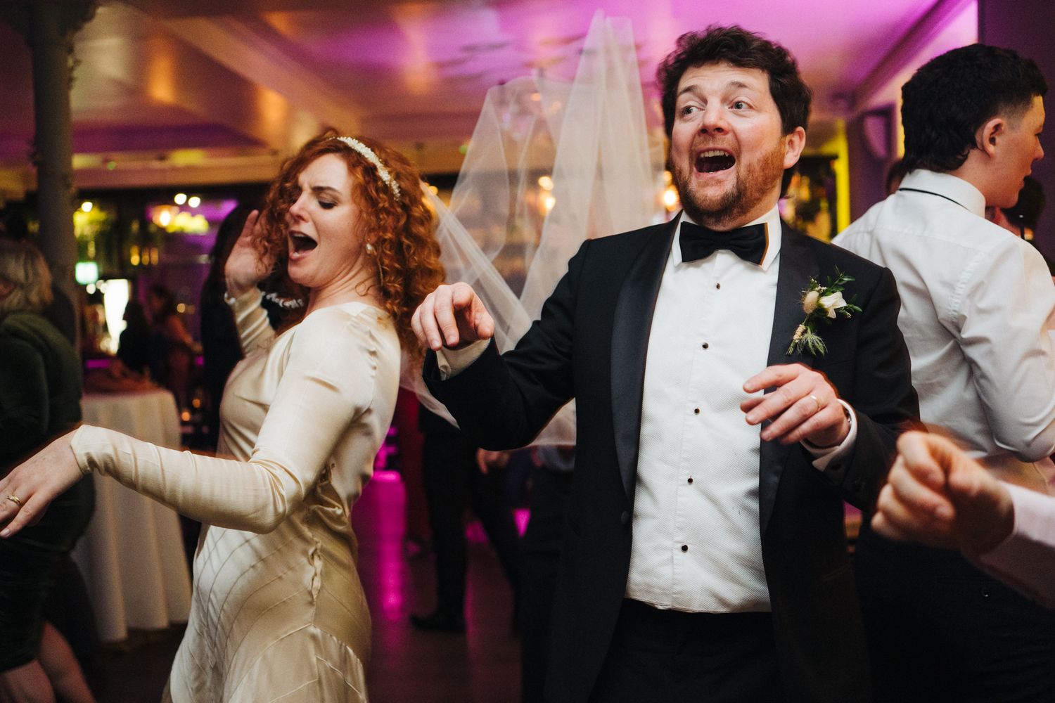 Wedding couple dancing enthusiastically at their reception under purple lighting.