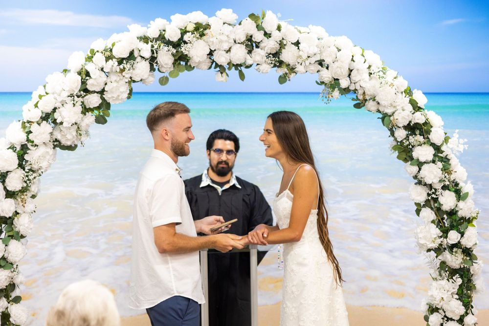 Intimate beach wedding ceremony with white floral arch and turquoise ocean backdrop.