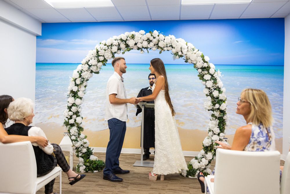 Wedding ceremony under floral arch with beach backdrop in indoor setting.