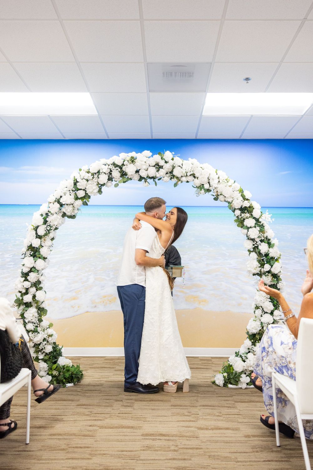 Romantic wedding moment under white floral arch with beach backdrop and seated guests.