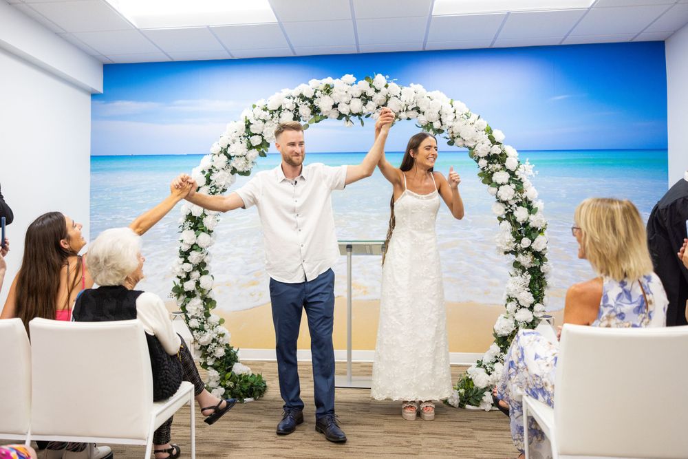 Wedding celebration with arms raised in joy under an elegant floral arch against ocean scenery.