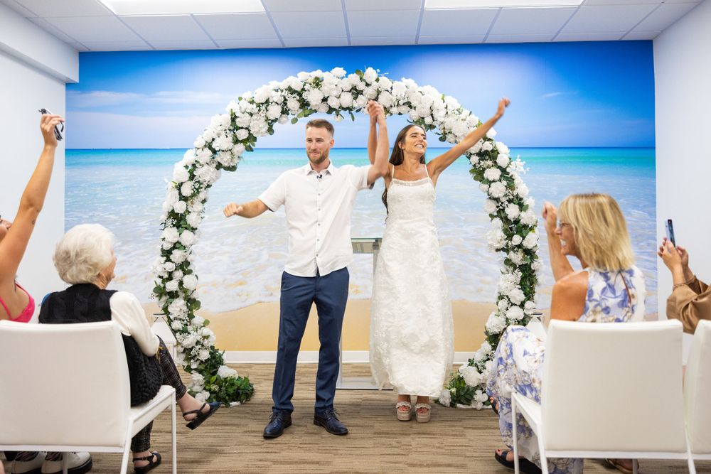 Newlyweds celebrate under a white floral arch with guests seated on white chairs beside a beach backdrop.