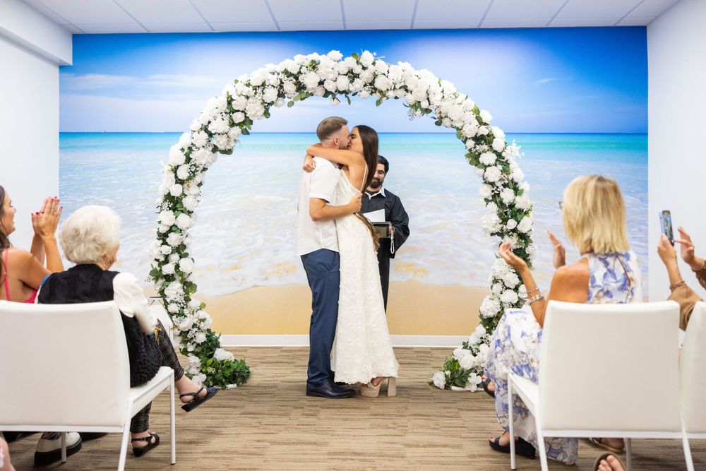 A joyful wedding ceremony takes place under a floral arch with white roses against a beach backdrop.