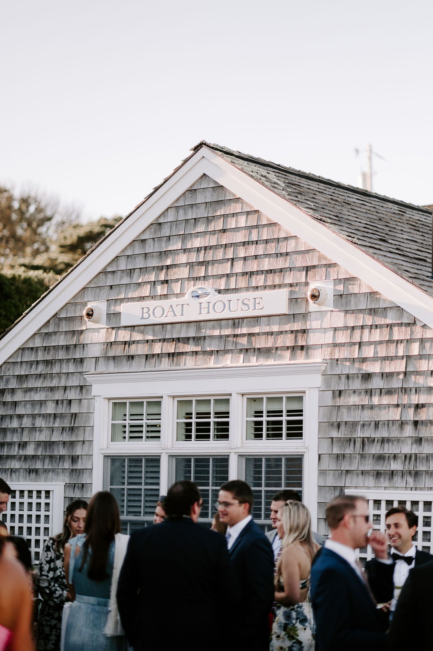 Wedding reception guests gather outside rustic boat house with weathered shingle siding.