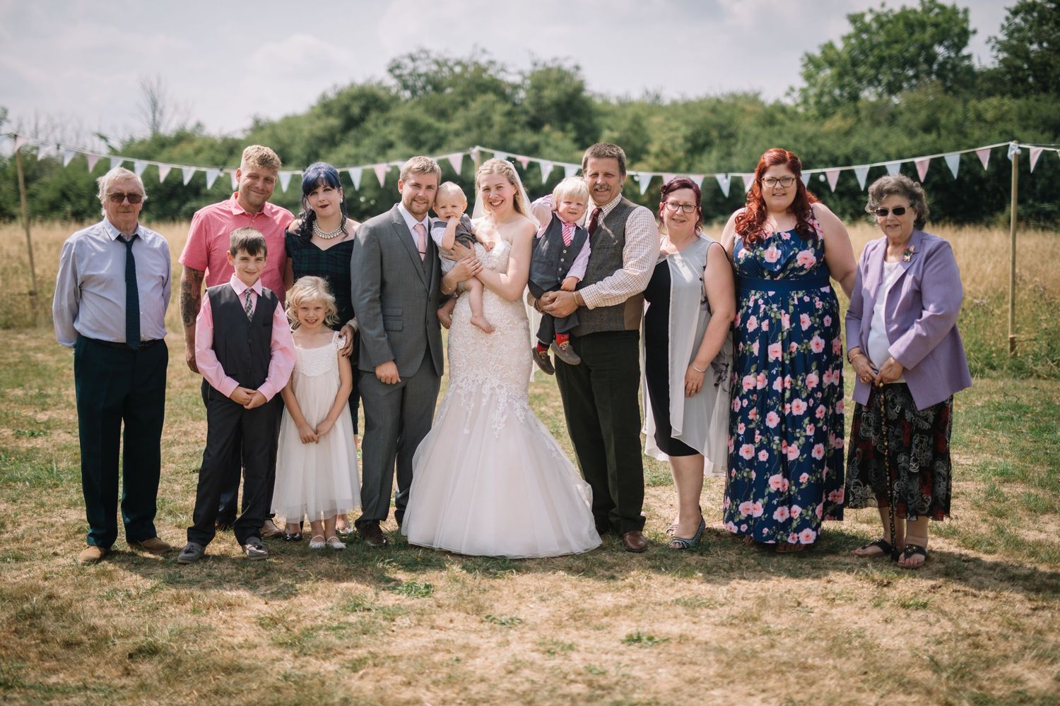 Wedding party group photo outdoors with bunting decorations and natural backdrop.
