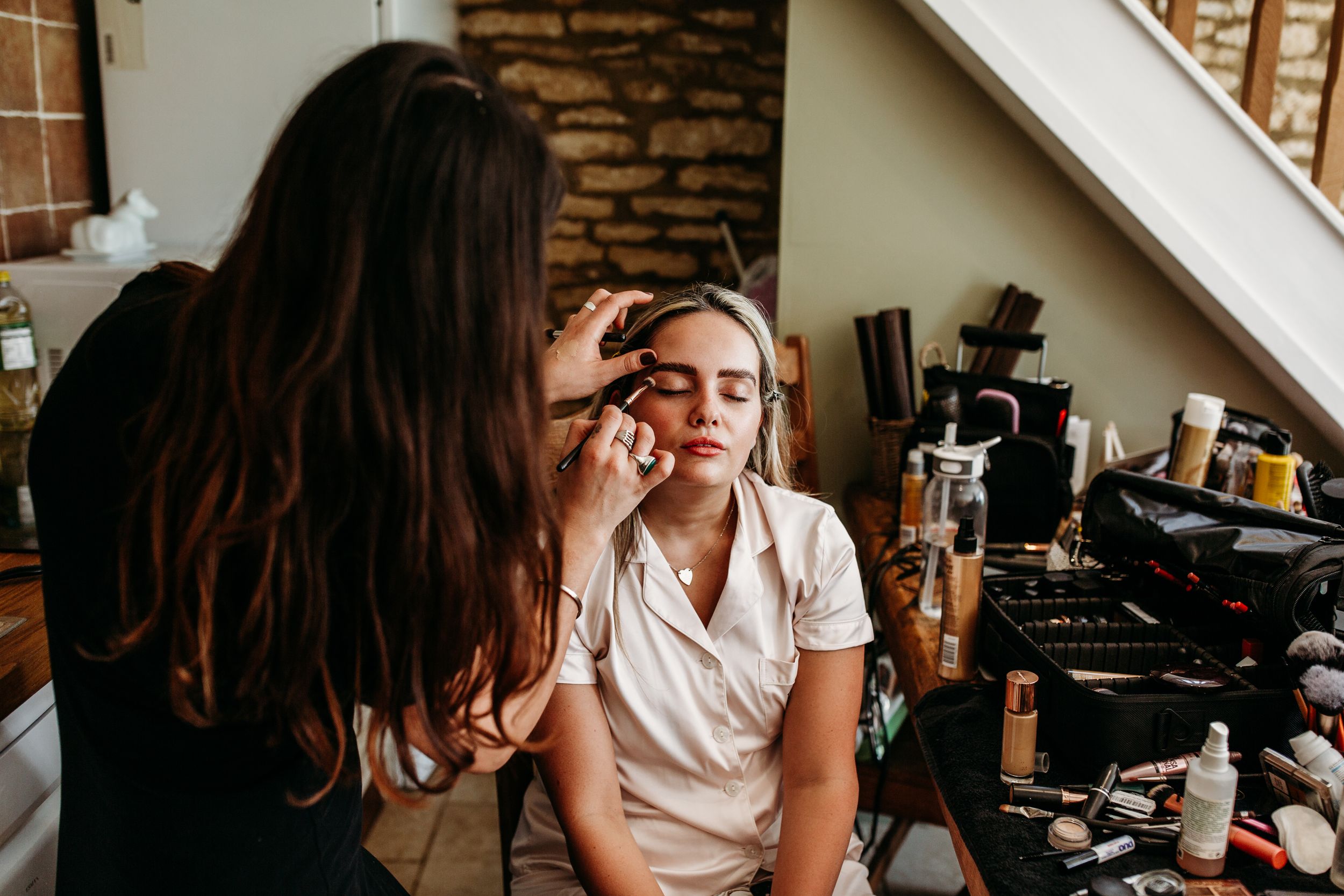 Makeup artist applying eye shadow to model during wedding day preparation in rustic venue.