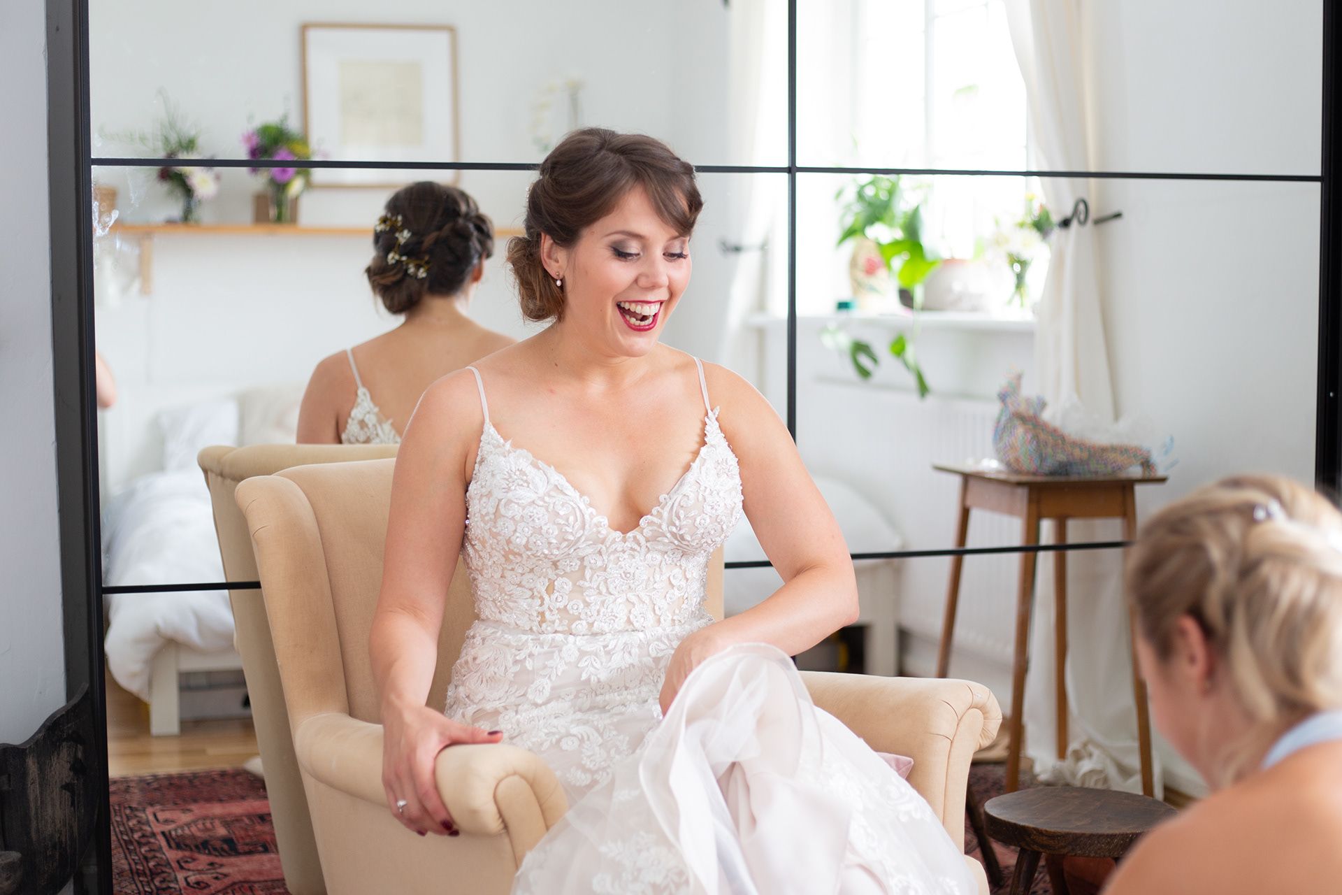 A beaming bride sits in a cream-colored chair wearing a delicate lace wedding dress.