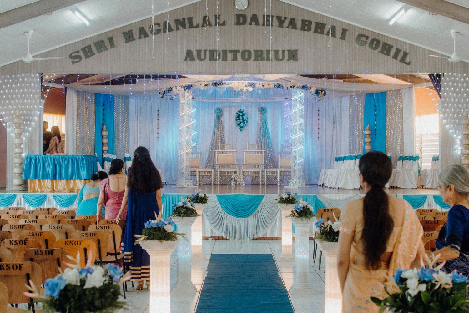 Wedding venue auditorium decorated with blue and white drapes, flowers, and chairs for ceremony.