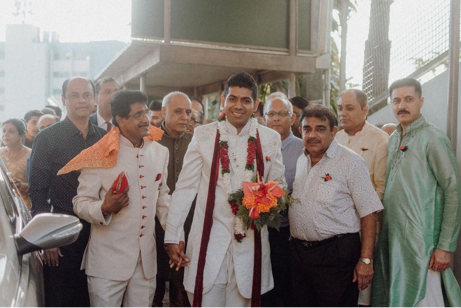 Wedding party and guests gather outside for group photo with groom in traditional Indian attire.