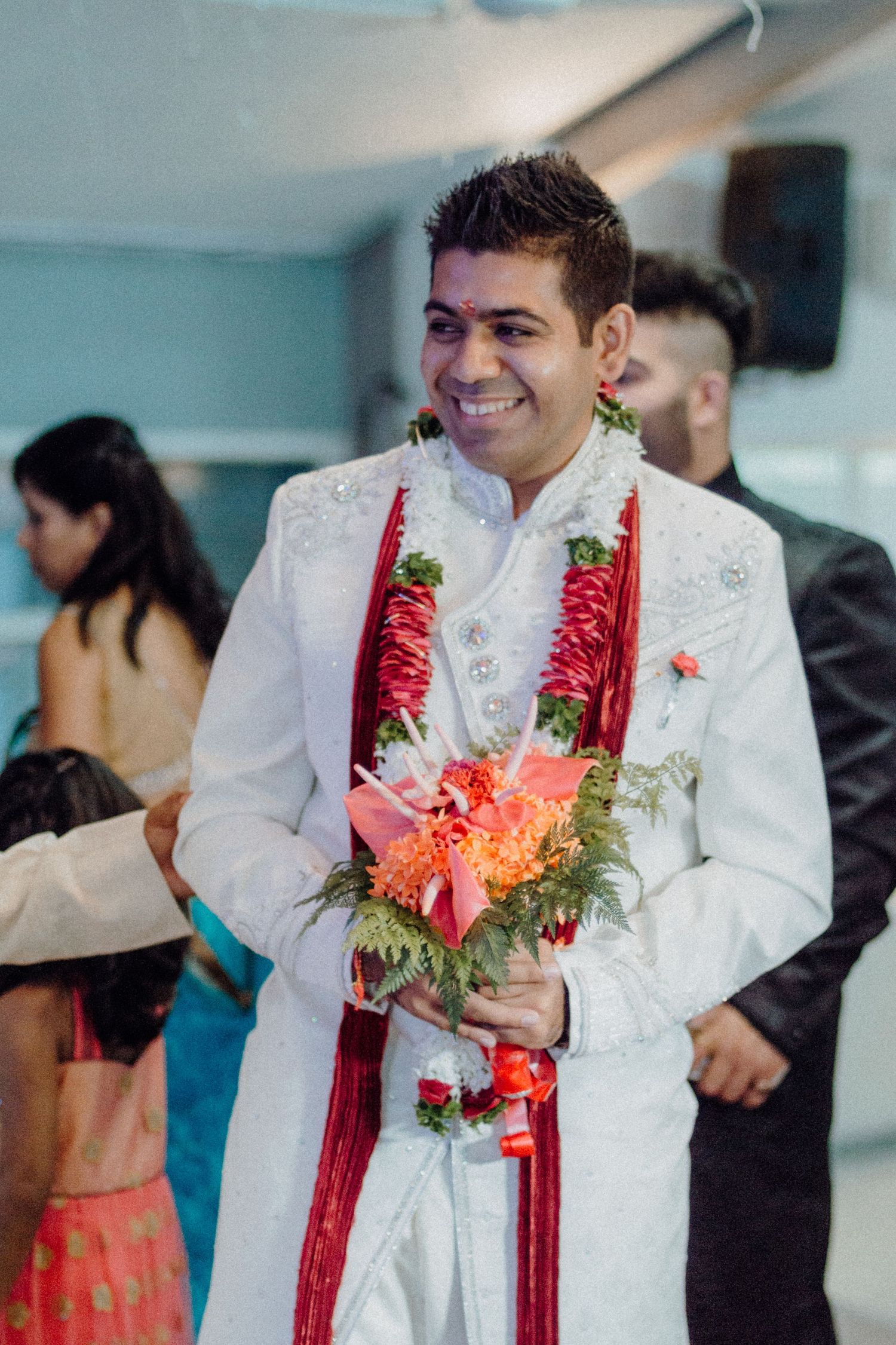 Groom in white sherwani with red garlands holding colorful flowers at Indian wedding.