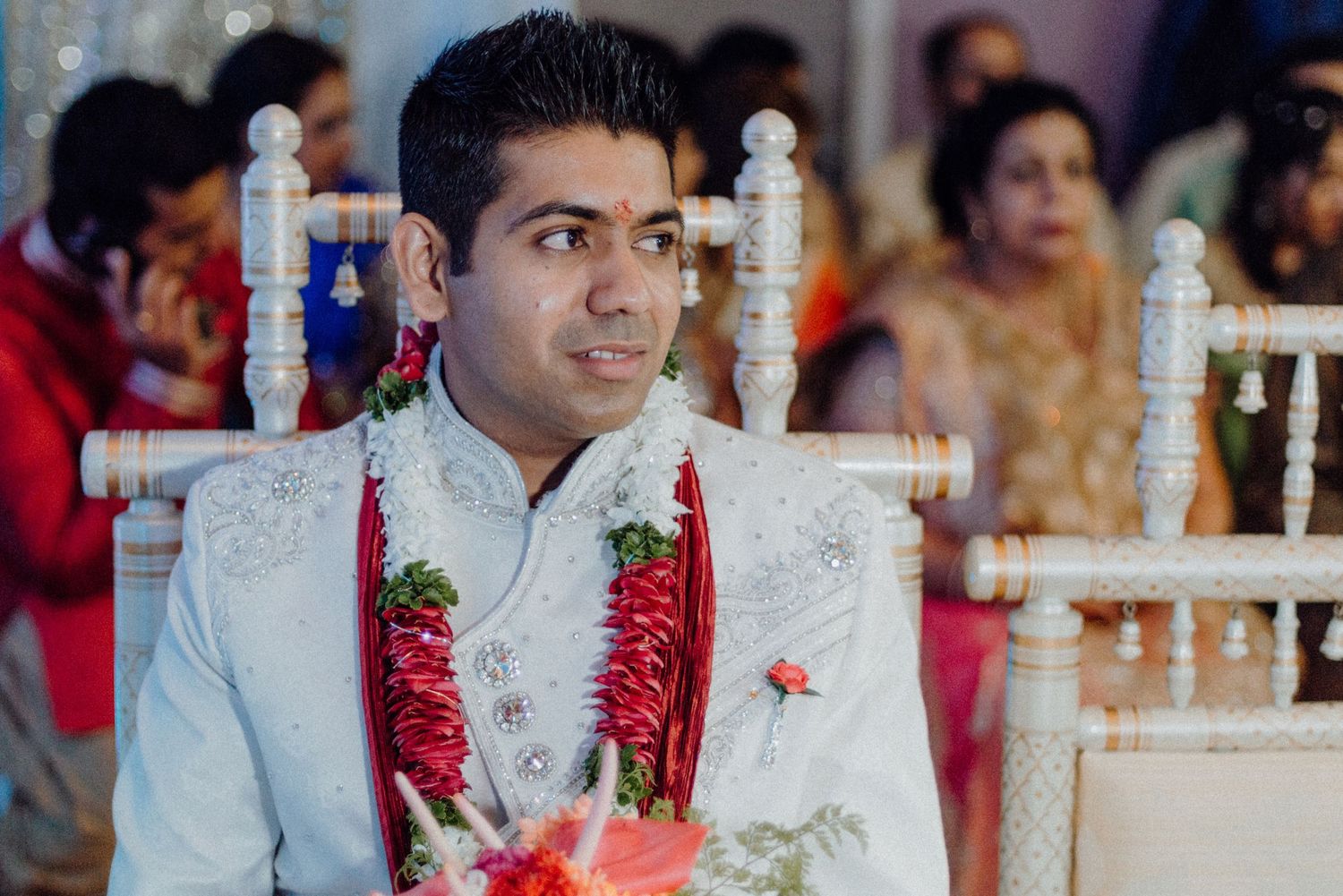 Groom in white sherwani with red garlands seated at decorated wedding venue.