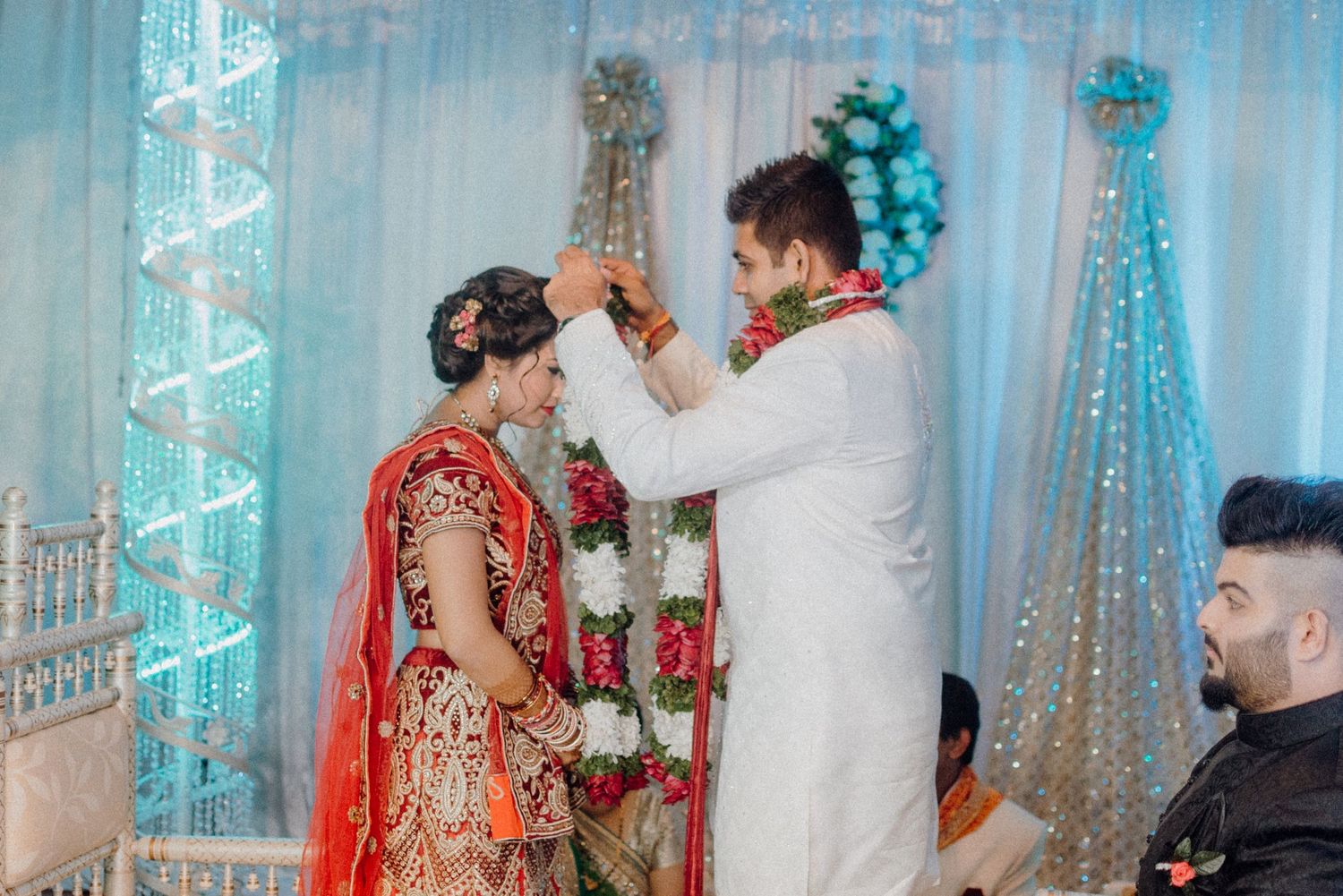 Wedding ceremony with couple exchanging garlands under blue and white decorations.