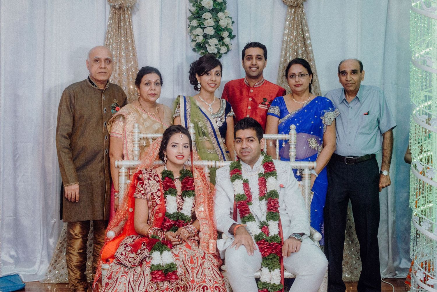Wedding portrait showing a couple in traditional Indian attire with family members against a decorated backdrop.