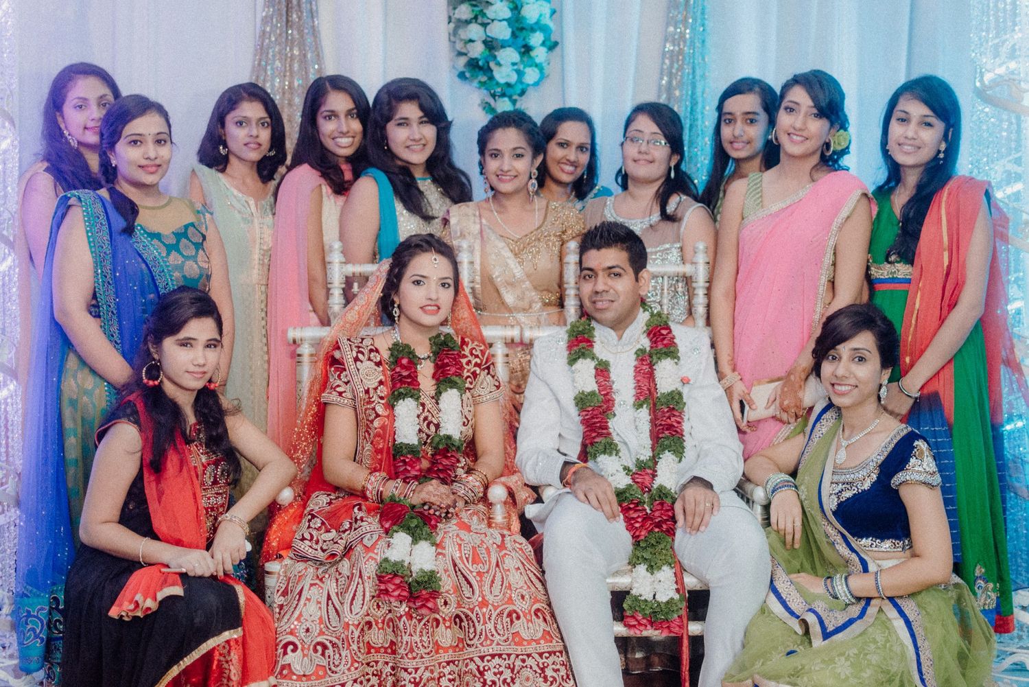 Wedding party photo featuring bridesmaids in colorful saris surrounding the couple in traditional ceremonial attire.
