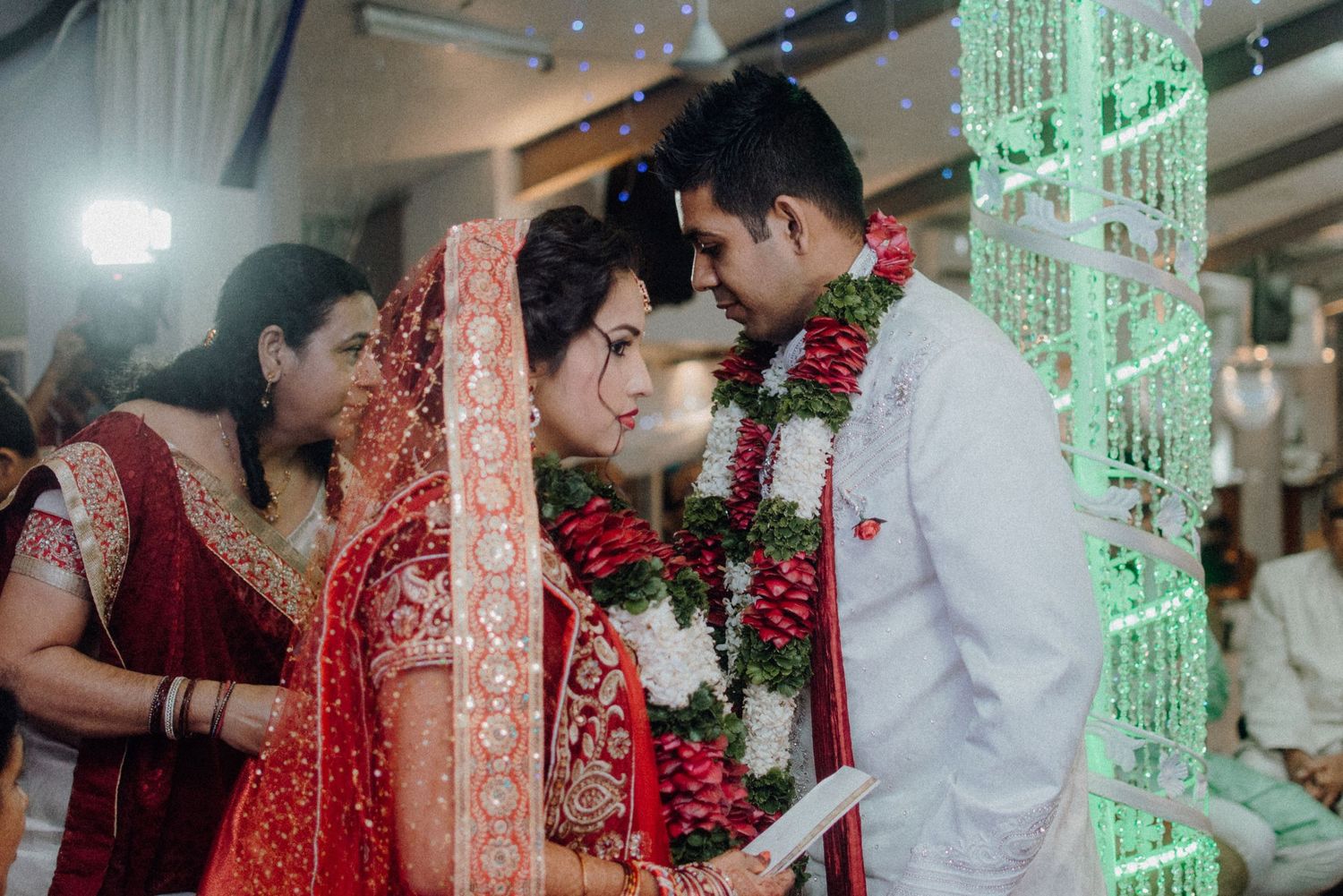 Intimate moment during an Indian wedding ceremony with decorative green lighting and floral garlands.