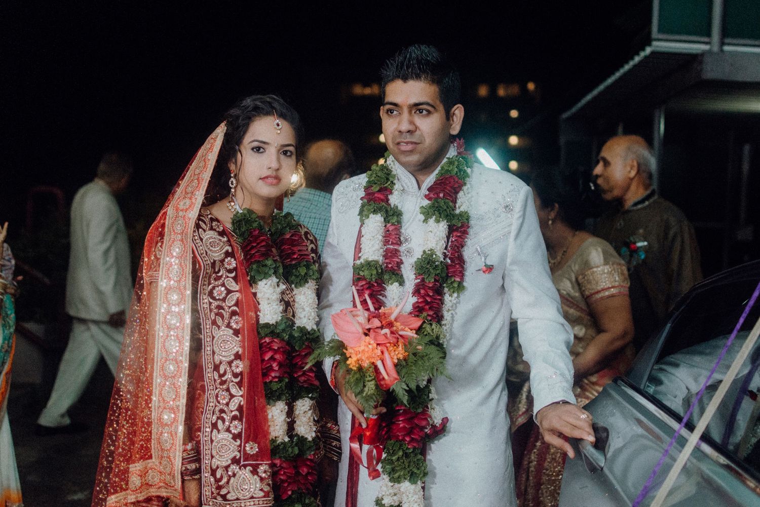 Newlyweds in traditional red and white wedding attire with floral garlands at night.