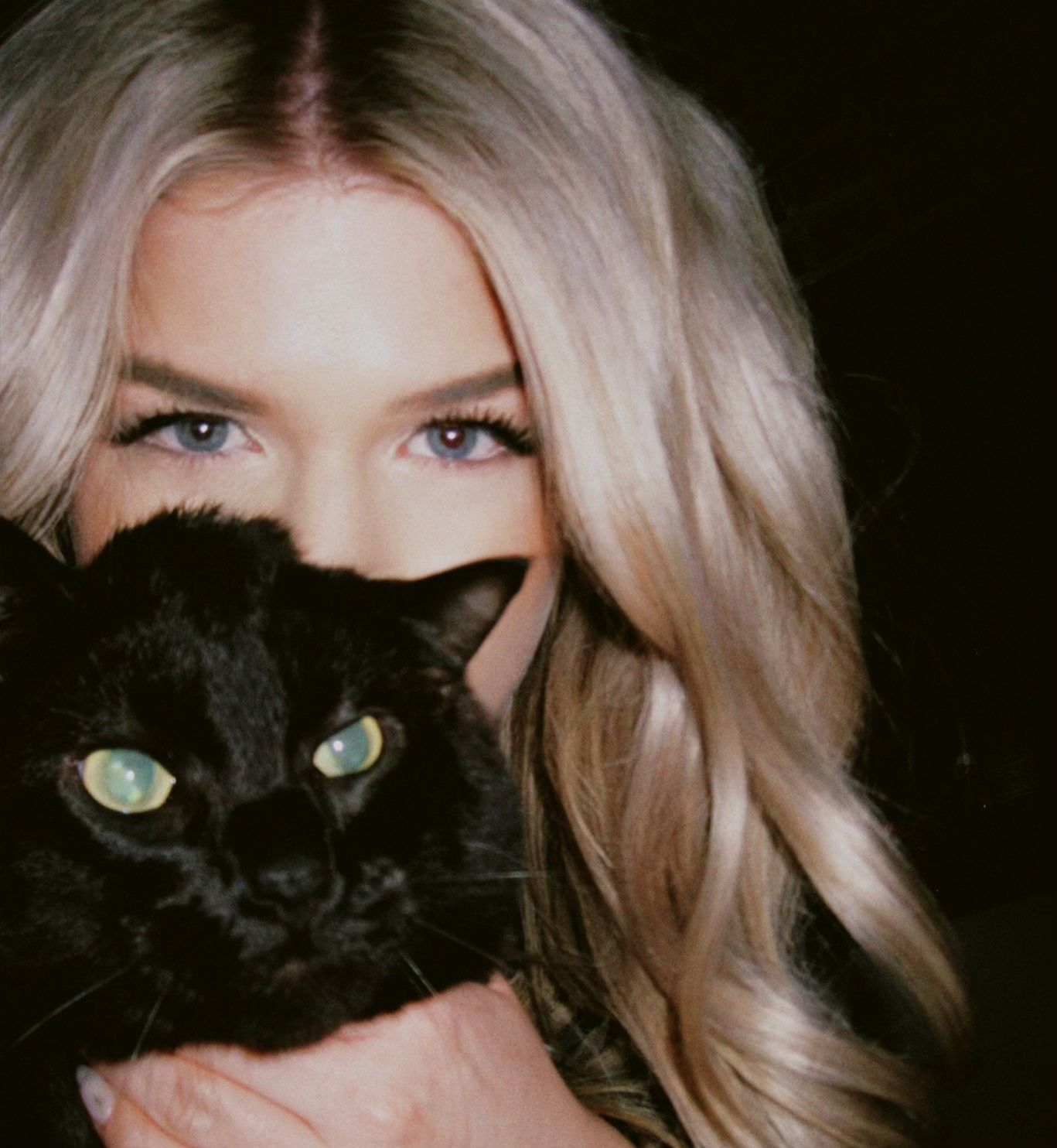 Blonde woman with blue eyes holds fluffy black cat close to her face in dark intimate portrait.