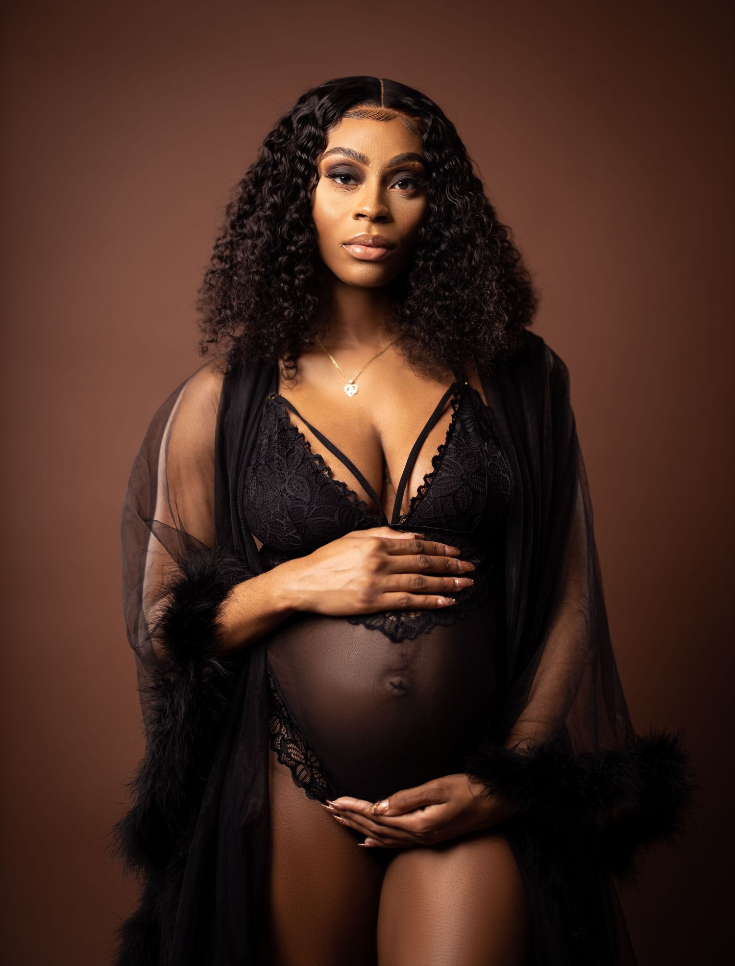 Maternity boudoir photo of woman in black lace lingerie against brown backdrop.