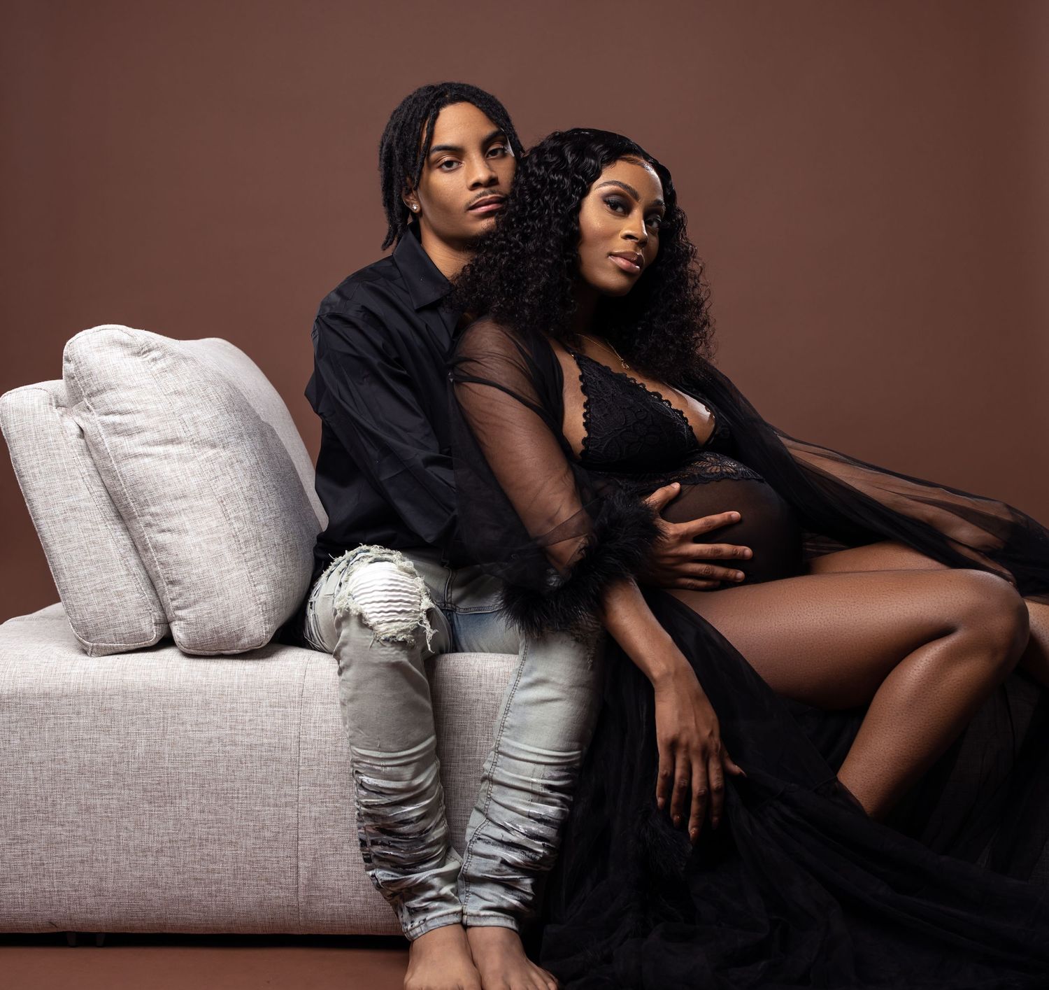 Intimate maternity portrait of couple lounging on gray sofa against brown background.