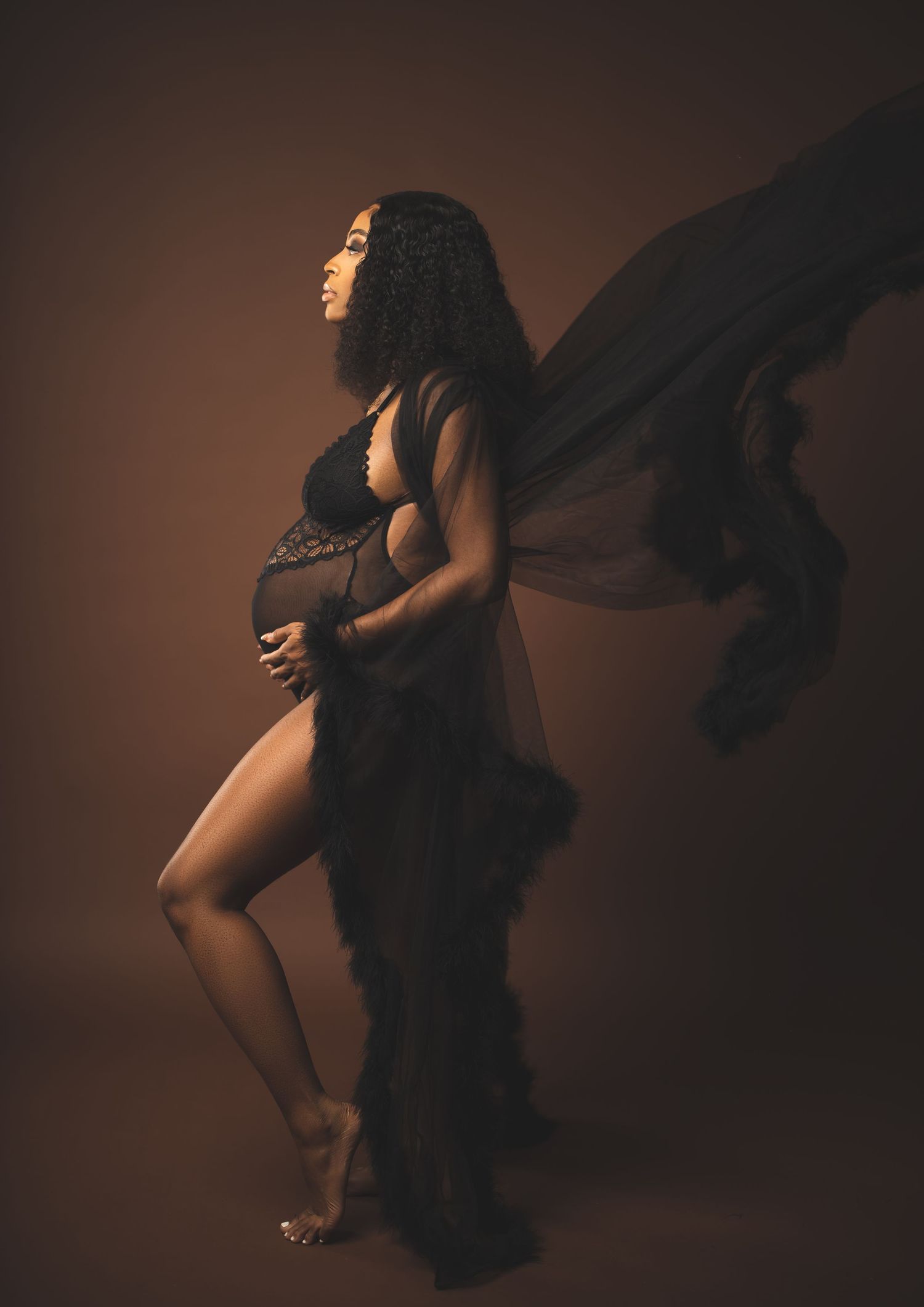 Artistic maternity silhouette with flowing fabric against moody brown background.