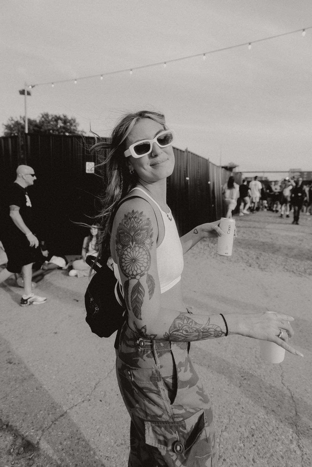 A tattooed person in sunglasses poses outdoors at a festival.