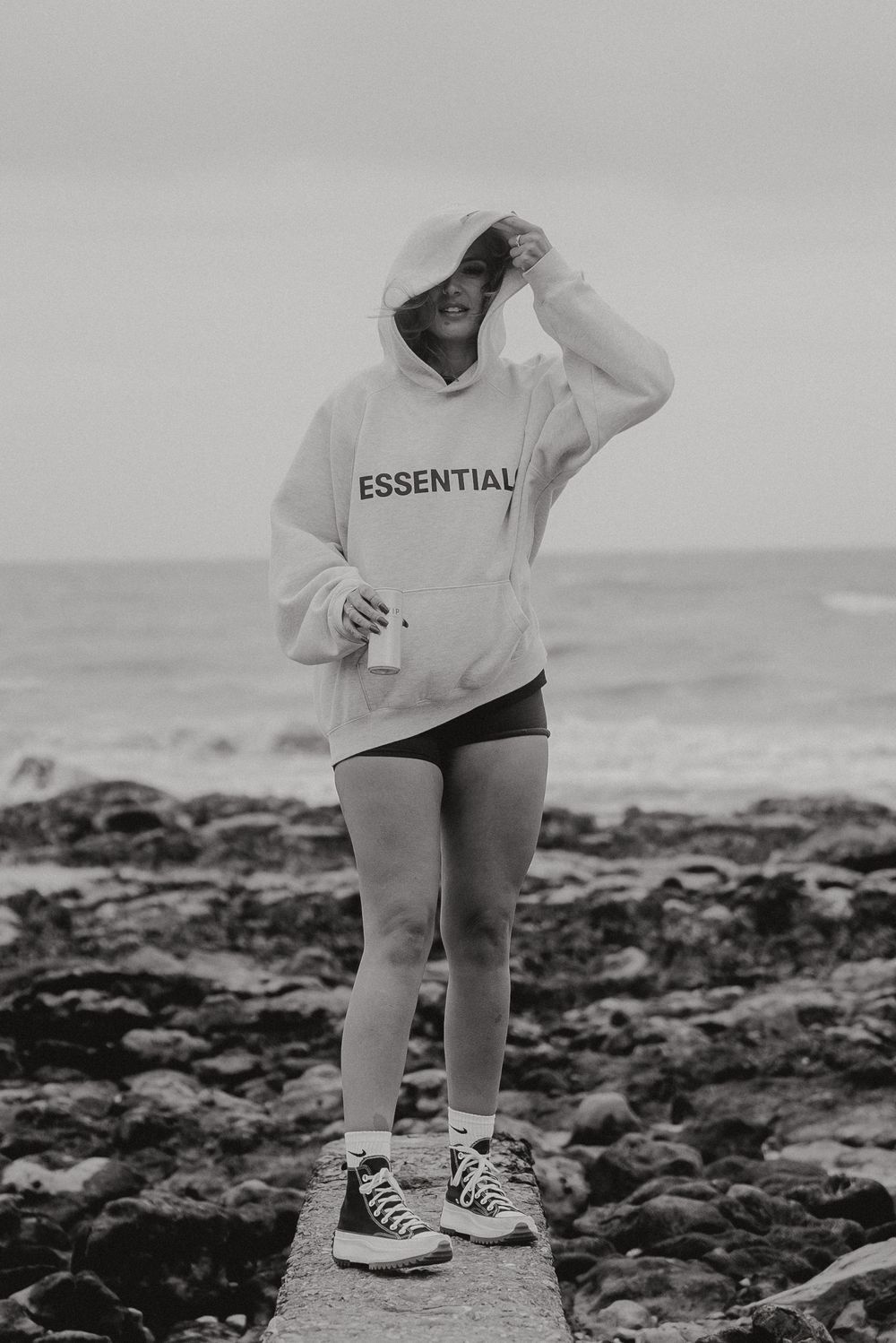 Person in 'Essential' hoodie standing on rocky coastline.