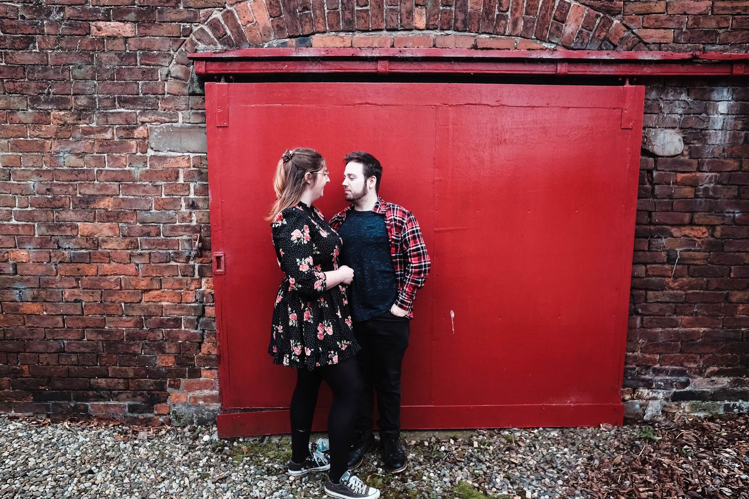 Natalie and Ryan leaning against a red farm house sliding door on their engagement shoot at Mill Bank Farm in Goostrey.