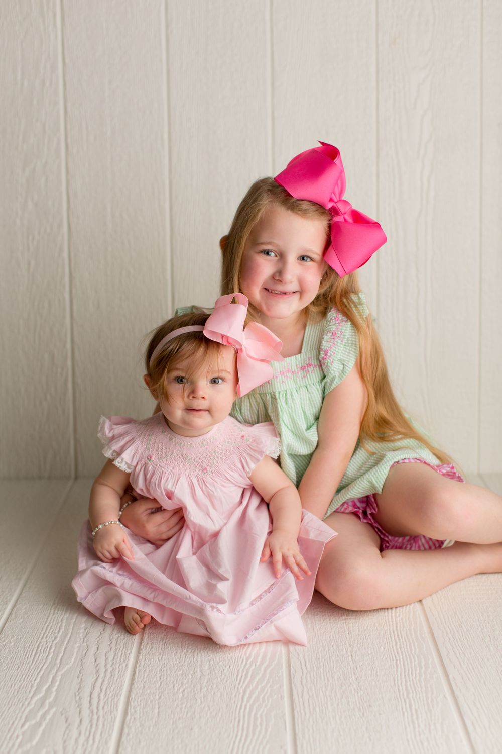 Two children in pastel dresses with pink bows posing together on white floor.