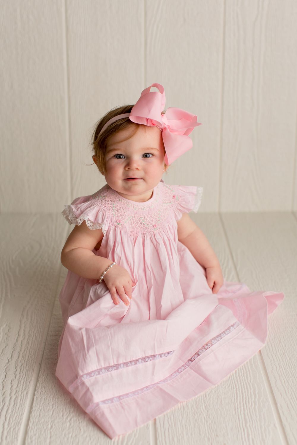 Small child in pink smock dress with lace trim and oversized bow sitting on white wooden floor.