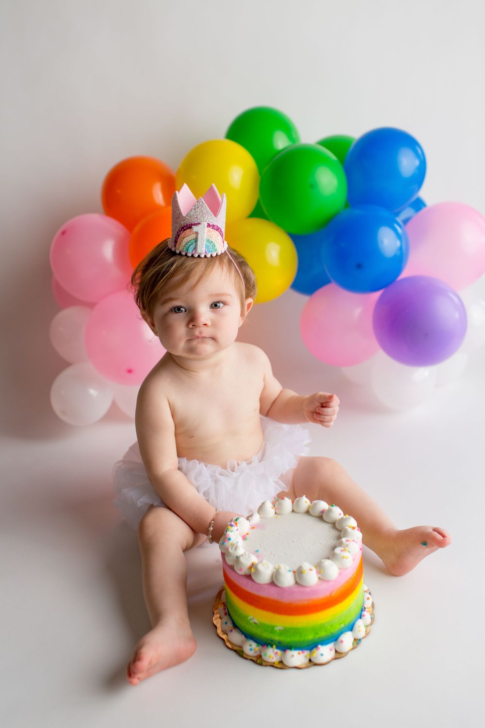 Baby in tutu with rainbow balloon arch and colorful birthday cake.