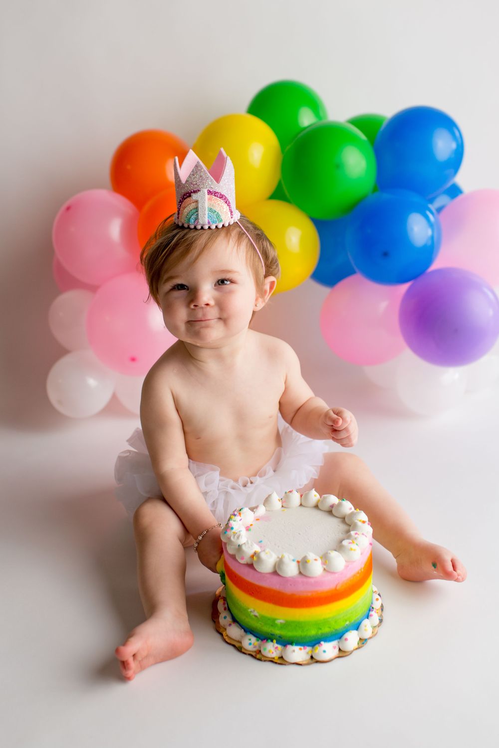 Colorful rainbow cake with whipped cream sits in front of balloon arch during festive birthday photoshoot.