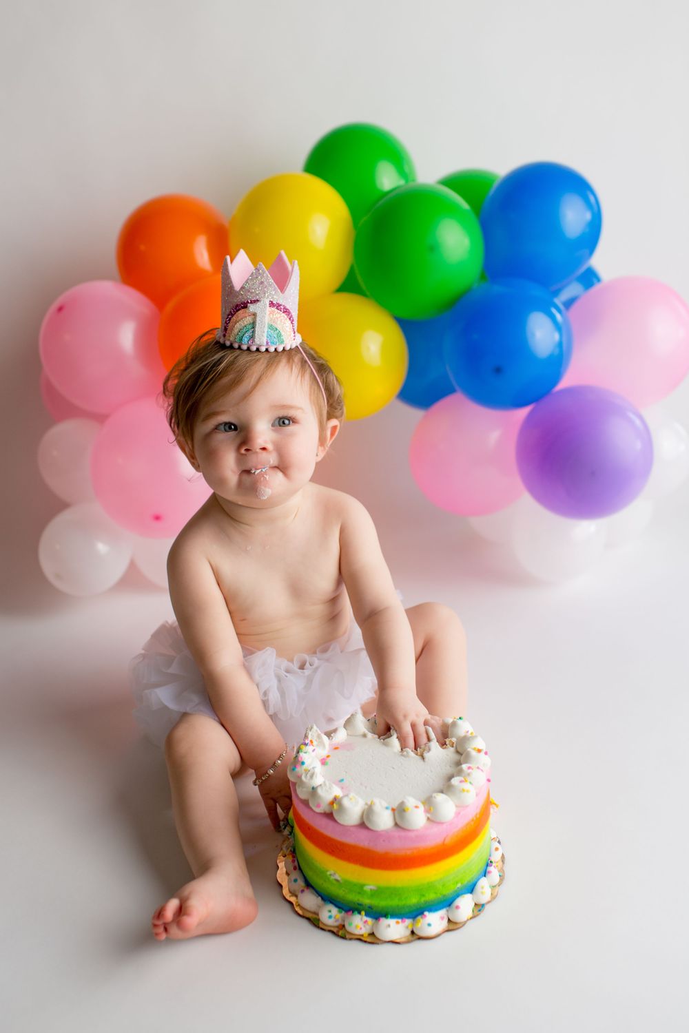 Rainbow birthday cake and multicolored balloon backdrop create cheerful celebration scene.