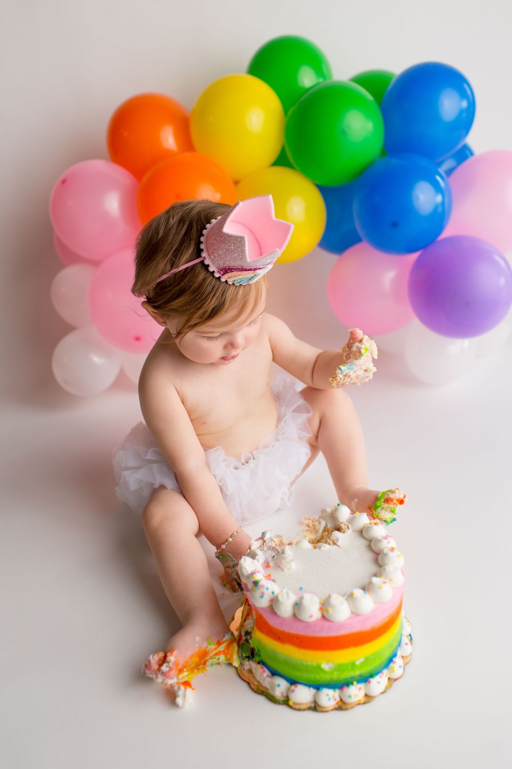 Smash cake session with rainbow cake and colorful balloon backdrop.