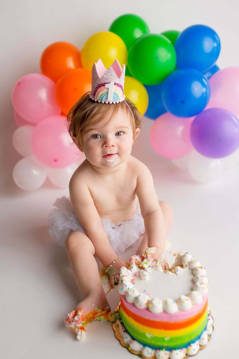 A rainbow cake smash photo featuring a unicorn crown, colorful balloons, and a tulle skirt.