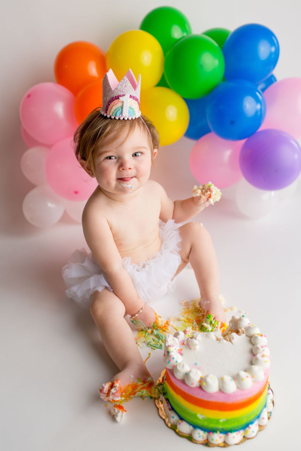 A festive cake smash session with rainbow balloons and a colorful layered cake.