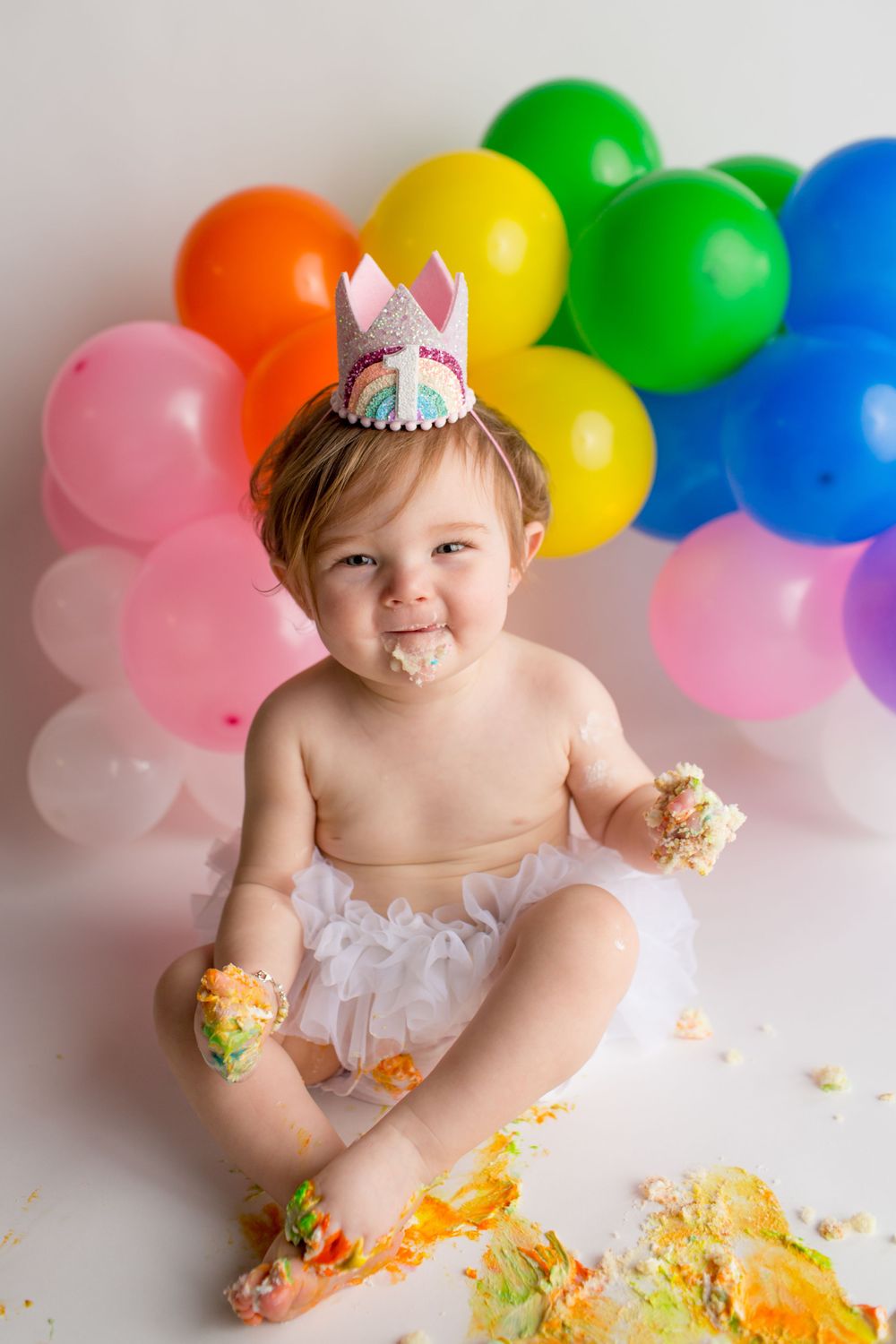 A messy cake smash photoshoot with rainbow balloons and scattered cake crumbs.