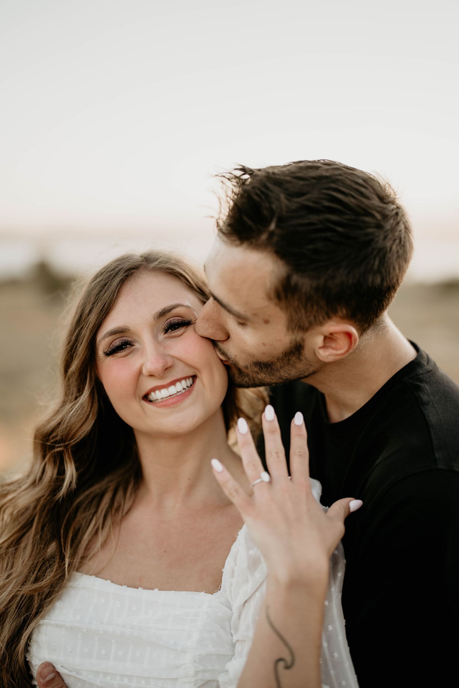 Newly engaged white couple flashing brand new engagement ring as they share a kiss on the cheek at Discovery Park WA