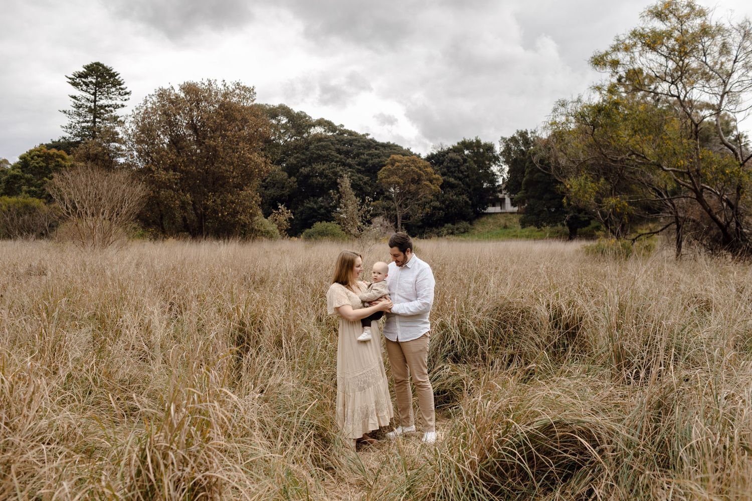 Couple holding baby in a golden grass field during an overcast family portrait session.