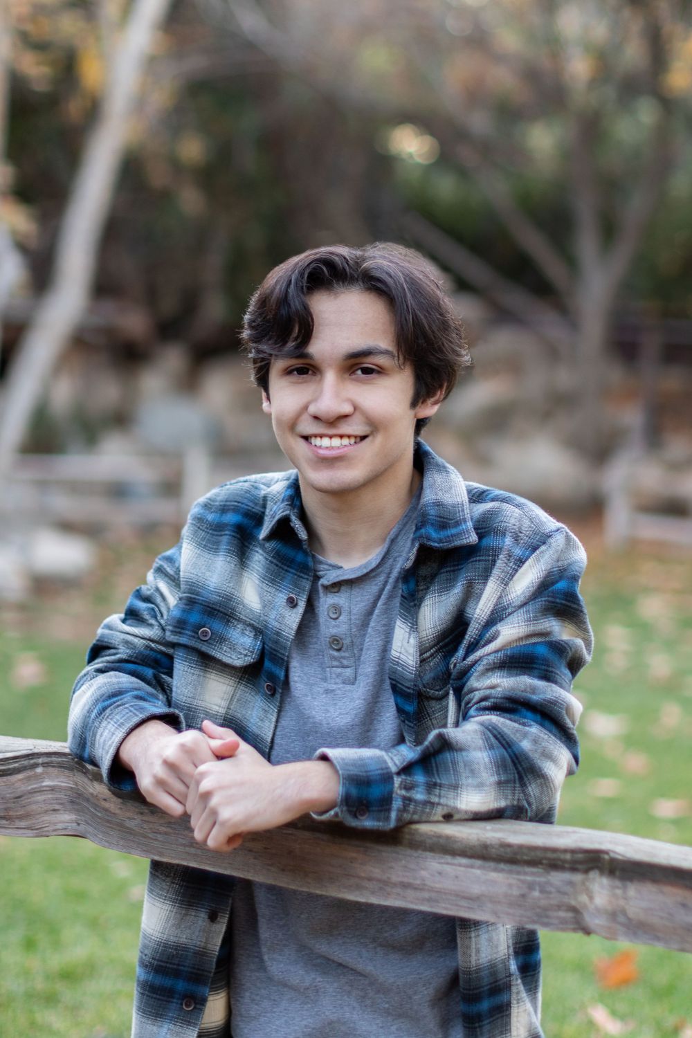 Orange County photographer captures  teenage boy in blue plaid flannel shirt leaning on a wooden fence in an autumn park setting.