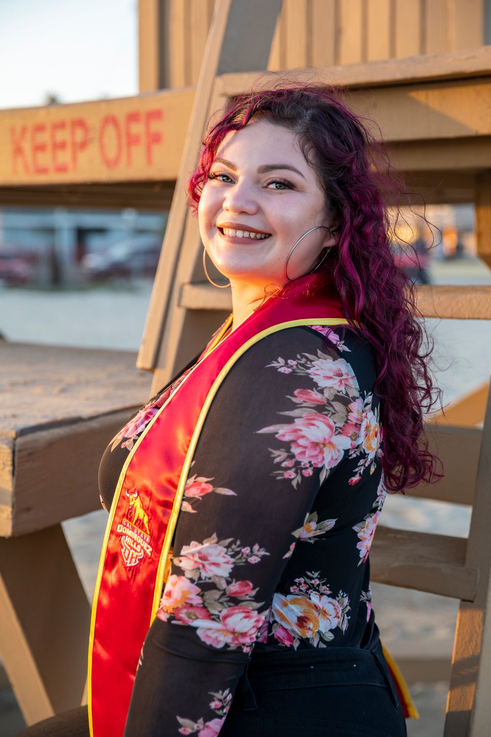 Orange County photographer captures Smiling graduate with purple hair wearing red and gold stole poses near a wooden lifeguard stand.