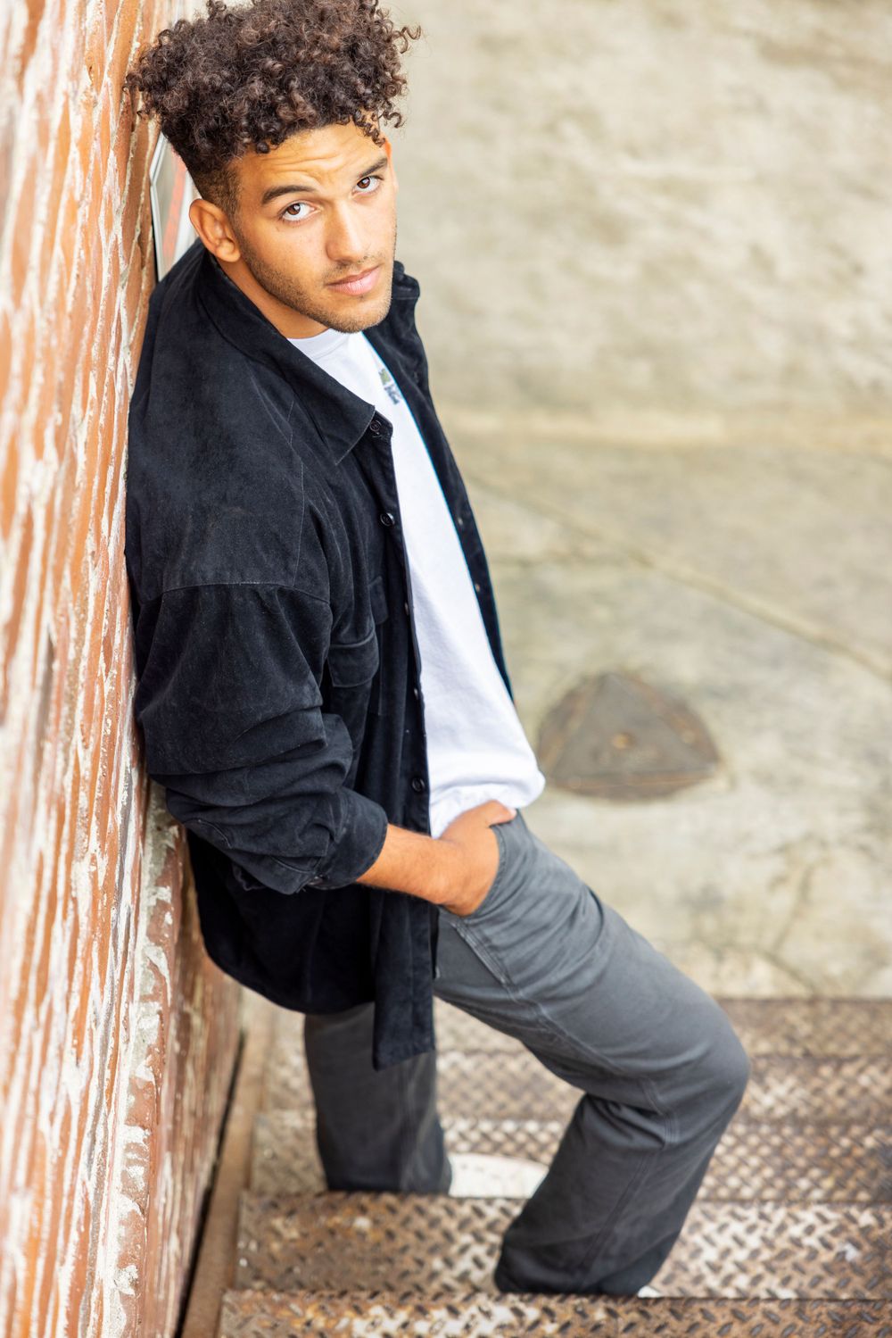 Orange County photographer captures Stylish young man in black jacket leaning against rustic wall on metal staircase with confident pose.