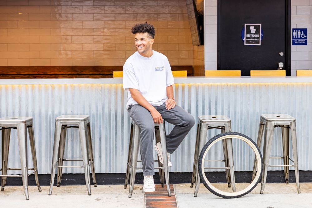 Orange County photographer captures Young man in white t-shirt sitting casually on metal barstool smiling in modern indoor venue.