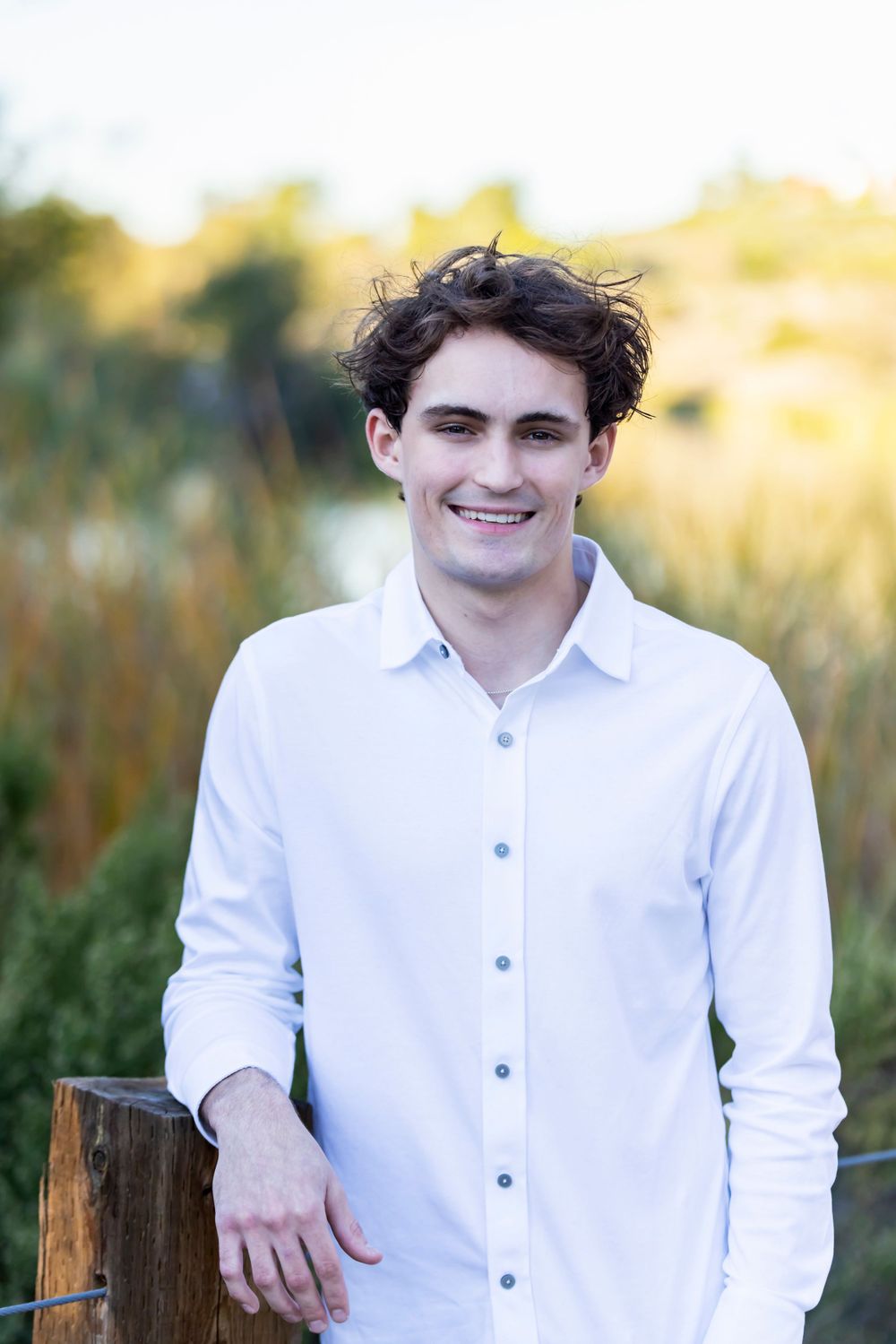 Orange County photographer captures Smiling young man in white button-up shirt leaning on wooden fence post in outdoor nature setting.