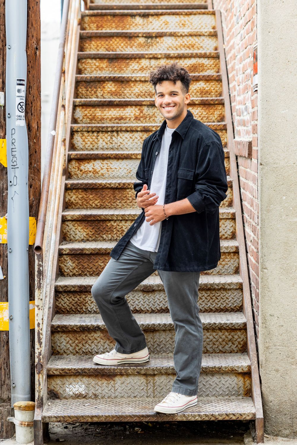 Orange County photographer captures Smiling young man in black jacket and gray pants posing on rustic metal outdoor staircase.