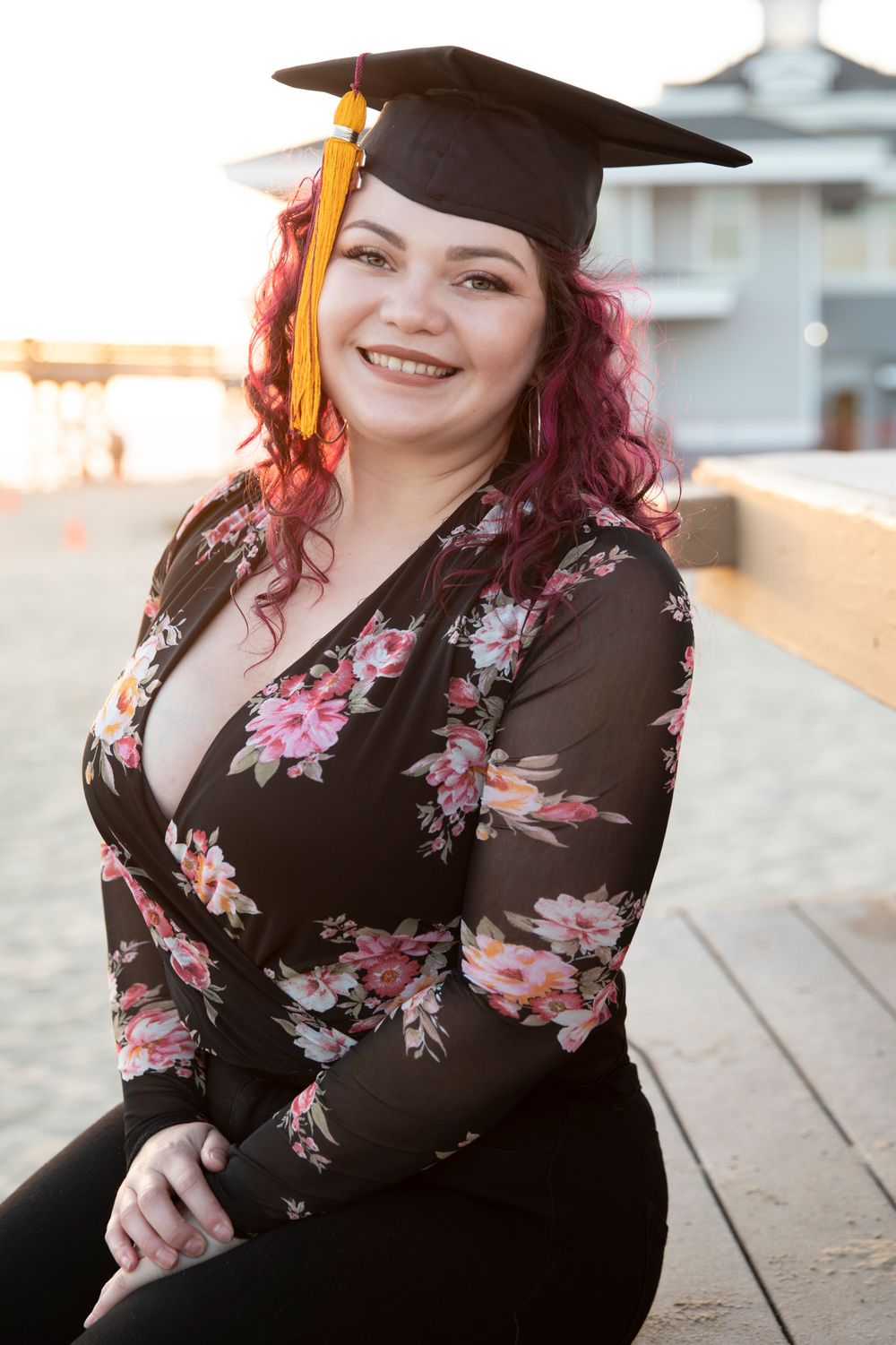 Orange County photographer captures Graduate with pink hair wearing mortarboard cap and floral top sits outdoors at sunset.