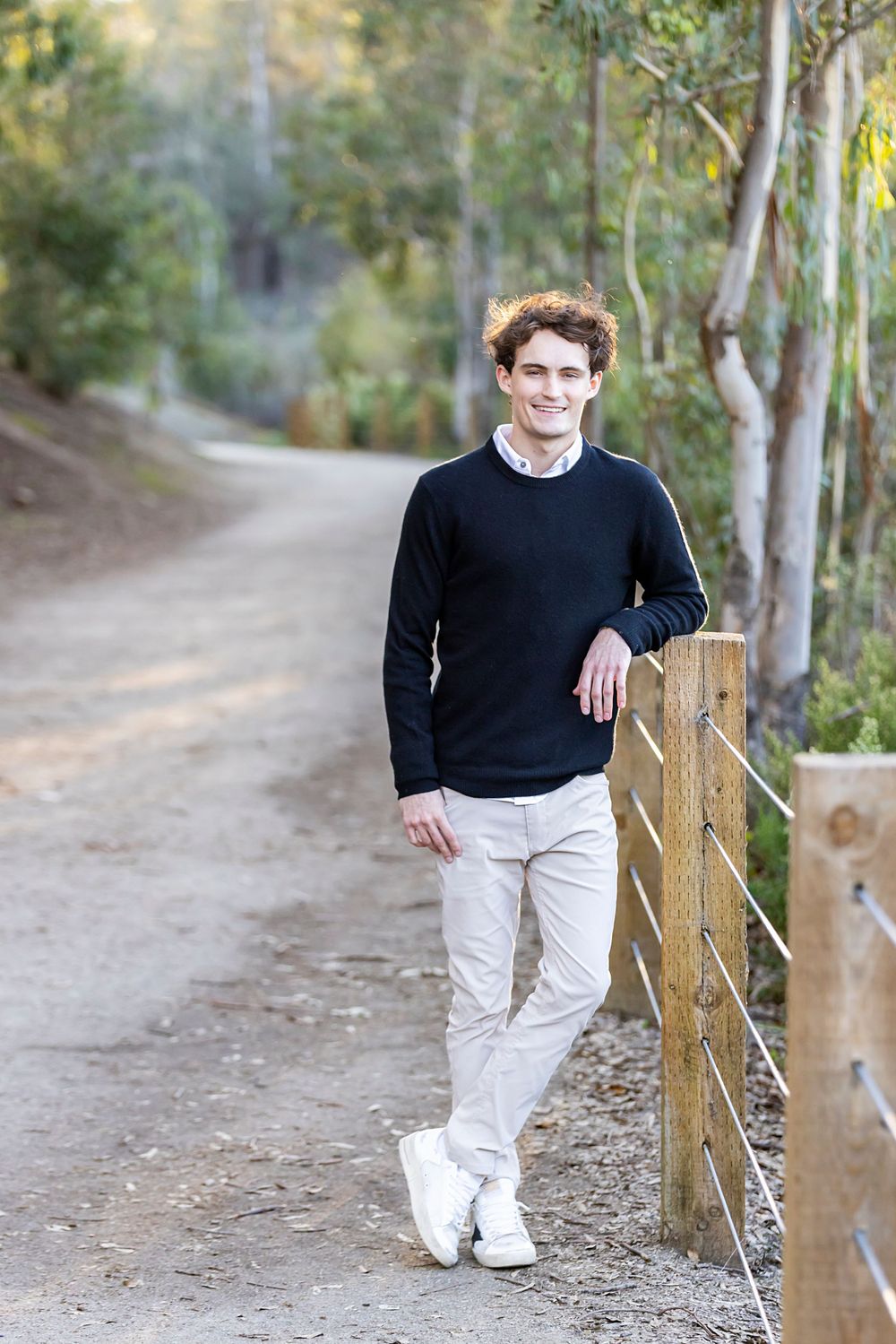 Orange County photographer captures Young man in black sweater and khakis leaning on wooden fence post along a tree-lined dirt path.