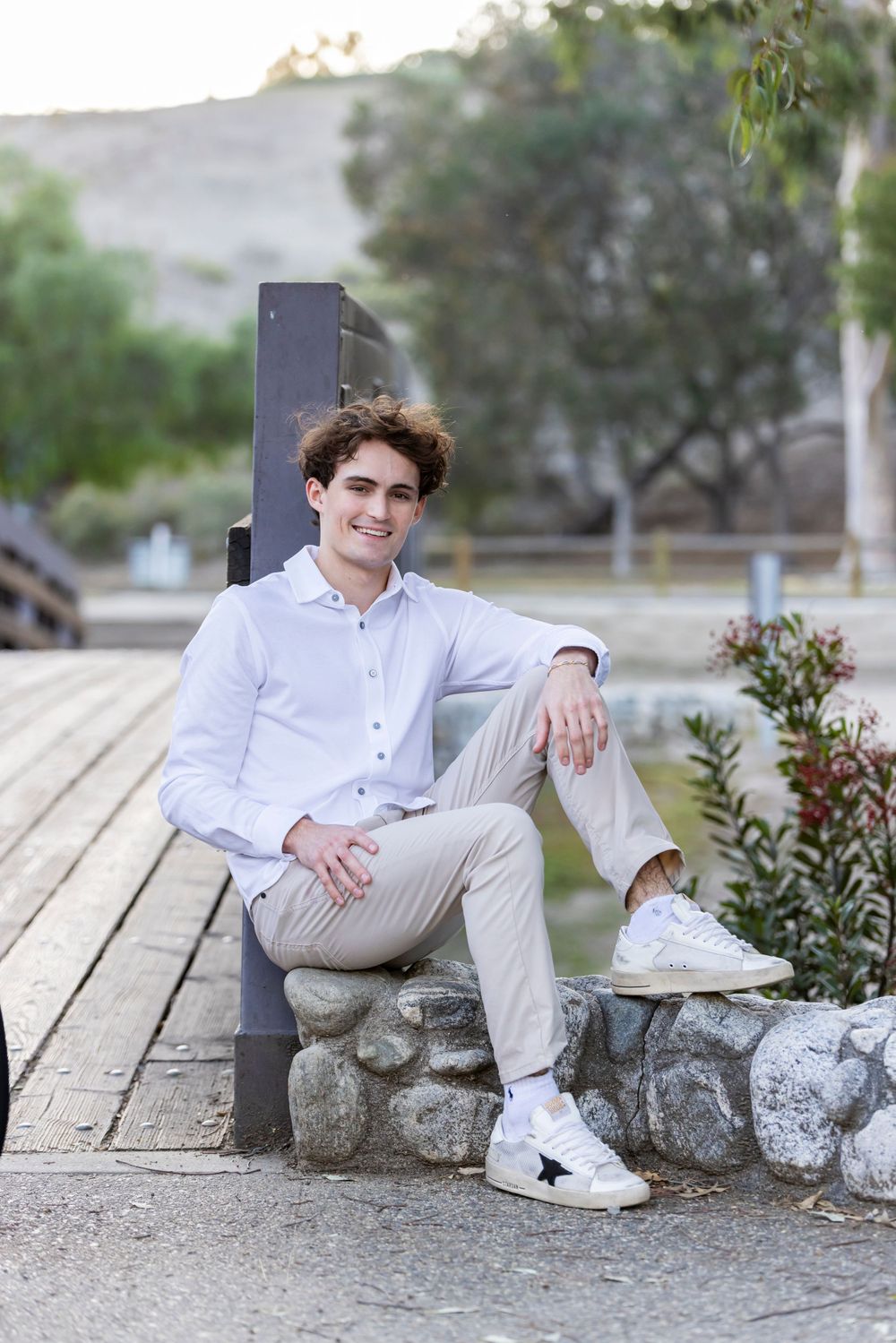 Orange County photographer captures Smiling young man in white shirt and khakis sitting relaxed on stone steps near a wooden bridge.