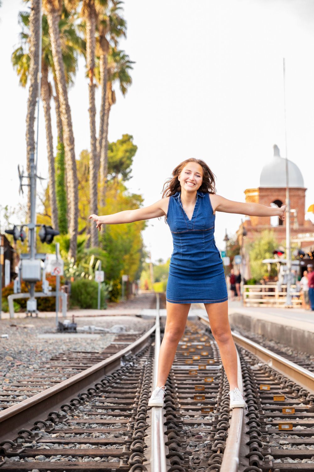 Orange County photographer captures young woman in blue dress standing on railroad tracks with arms outstretched near palm trees.