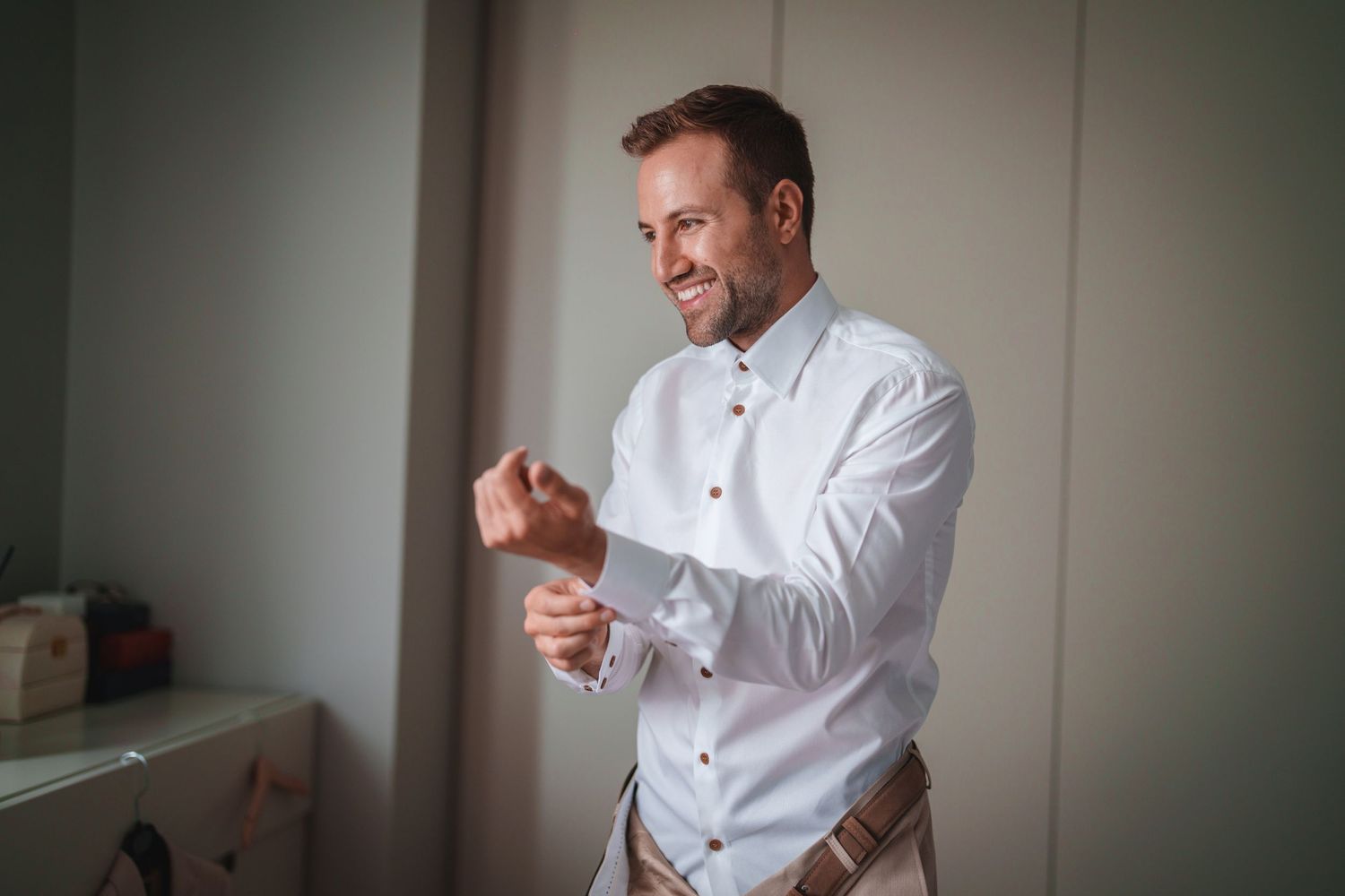 A person in white adjusts their cufflinks while smiling in a bedroom.
