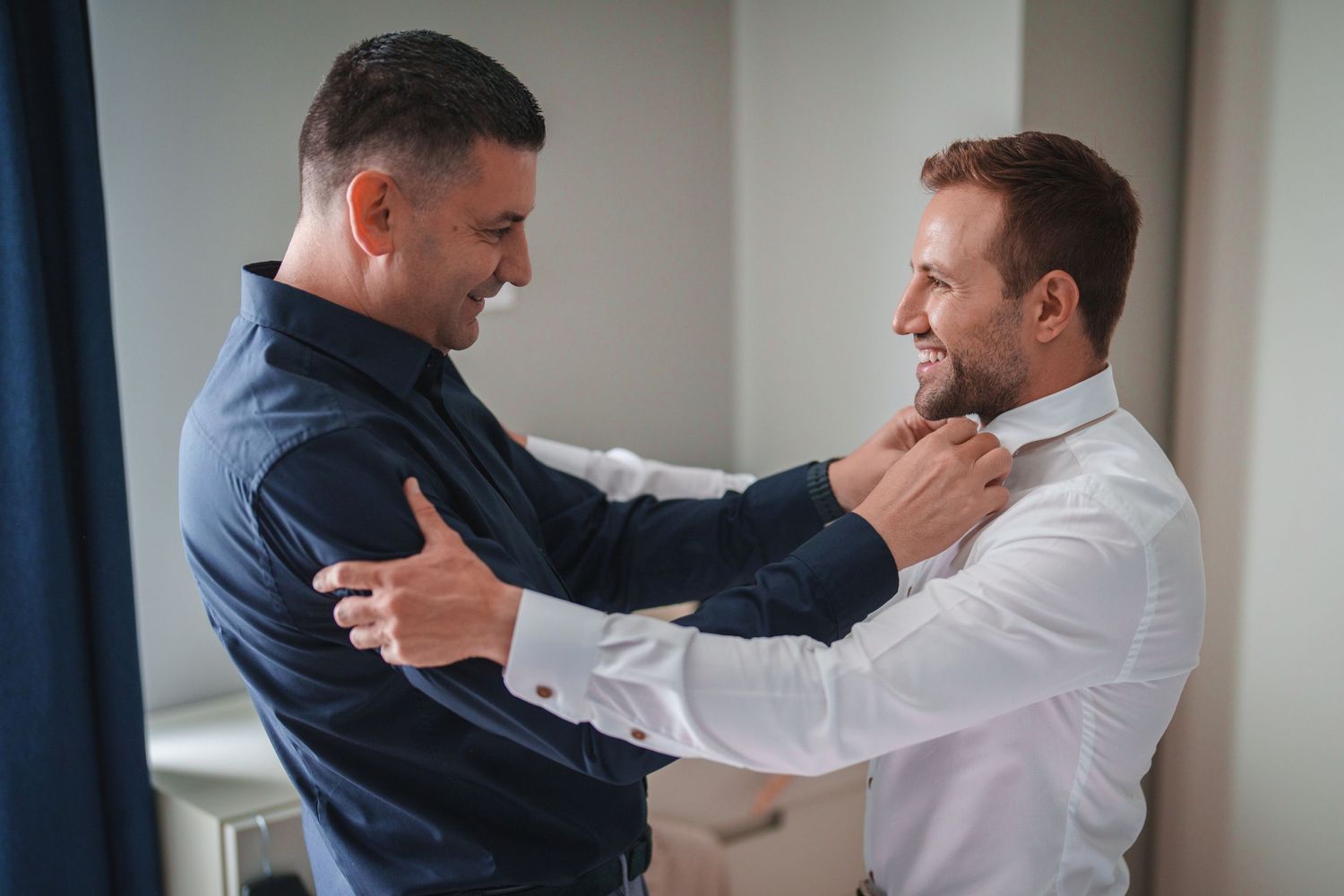 Two people help each other adjust their formal attire while sharing a laugh.